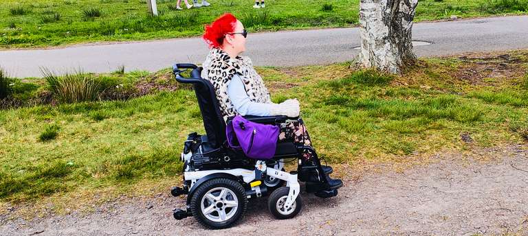 Side view of me in my wheelchair at Chasewater park, bright red hair standing out against the green grass. My trusty purple bag is hanging on the chair and I'm all cosy in my leopard print coat. The path's not too bad here - but the grass might be a different story! Accessibility inspector on duty!