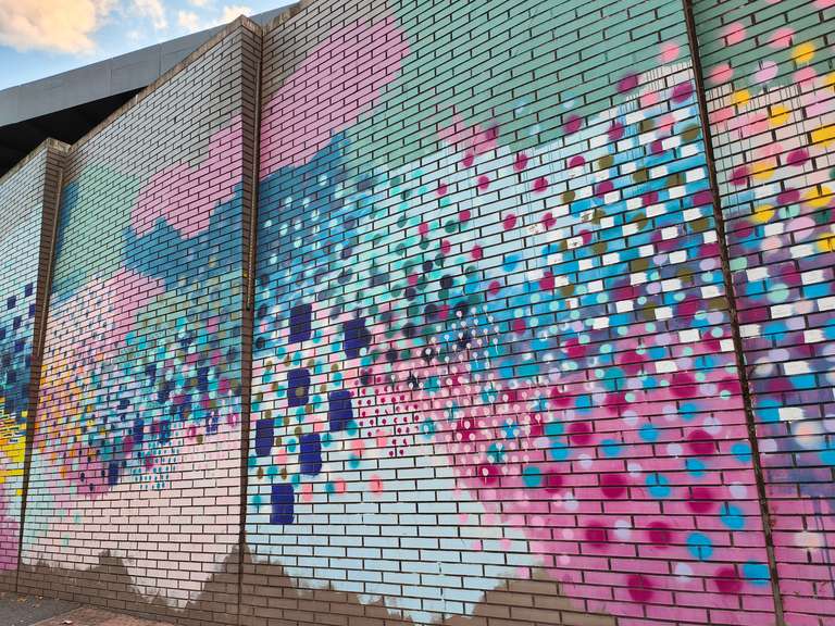 A vibrant brick wall painted with colourful abstract patterns in pastel pinks, blues, yellows, and purples. The mural creates a cheerful splash of colour against the otherwise plain building.