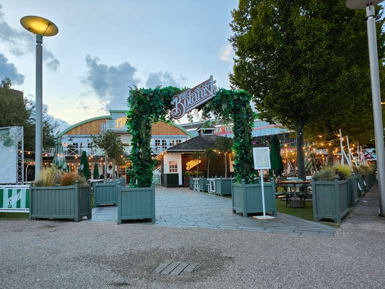 The entrance to The Botanist restaurant and bar at twilight, featuring a prominent green archway made of artificial foliage with the restaurant's logo sign. The path is paved and accessible, with outdoor seating areas visible.