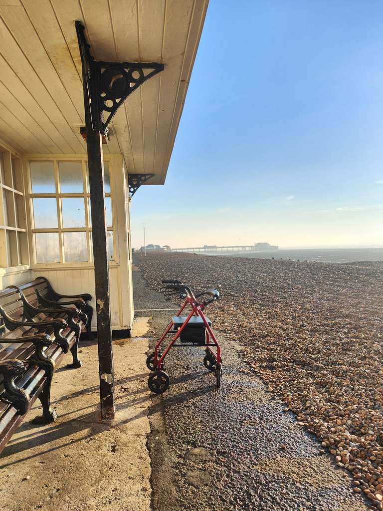 A vintage beach shelter on Brighton's pebble beach at golden hour, with a red mobility walker parked beneath it. In the distance, Brighton Pier can be seen along with the vast expanse of the English Channel.