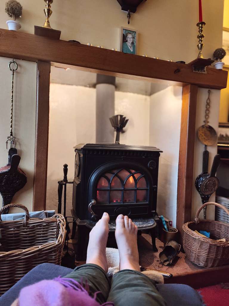 A cosy living room with feet propped up in front of a cast iron fireplace with glowing embers. Wicker baskets flank the hearth, and above the mantelpiece is a family photo. The scene suggests comfort and warmth.