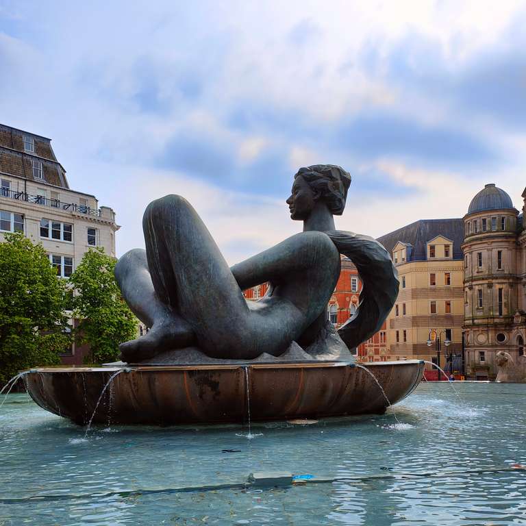 Birmingham's 'Floozy in the Jacuzzi' - A large bronze sculpture of a reclining female figure in a fountain in what appears to be Birmingham city centre. The graceful statue is positioned in a bowl-shaped basin with water features, surrounded by historic buildings.