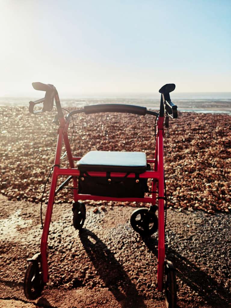 A bright red mobility walker sitting alone on a pebble beach, facing the sparkling sea with the silhouette of a pier in the distance. The walker casts a long shadow across the stones in the golden evening light.