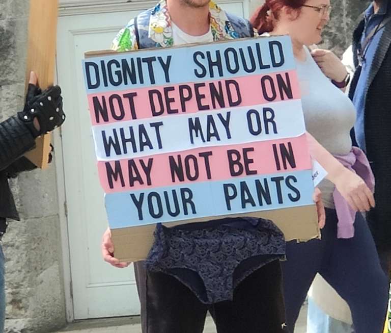 A protest sign with trans flag colours (blue, pink, white) reading 'DIGNITY SHOULD NOT DEPEND ON WHAT MAY OR MAY NOT BE IN YOUR PANTS', held by someone wearing a floral-patterned shirt at a rally.