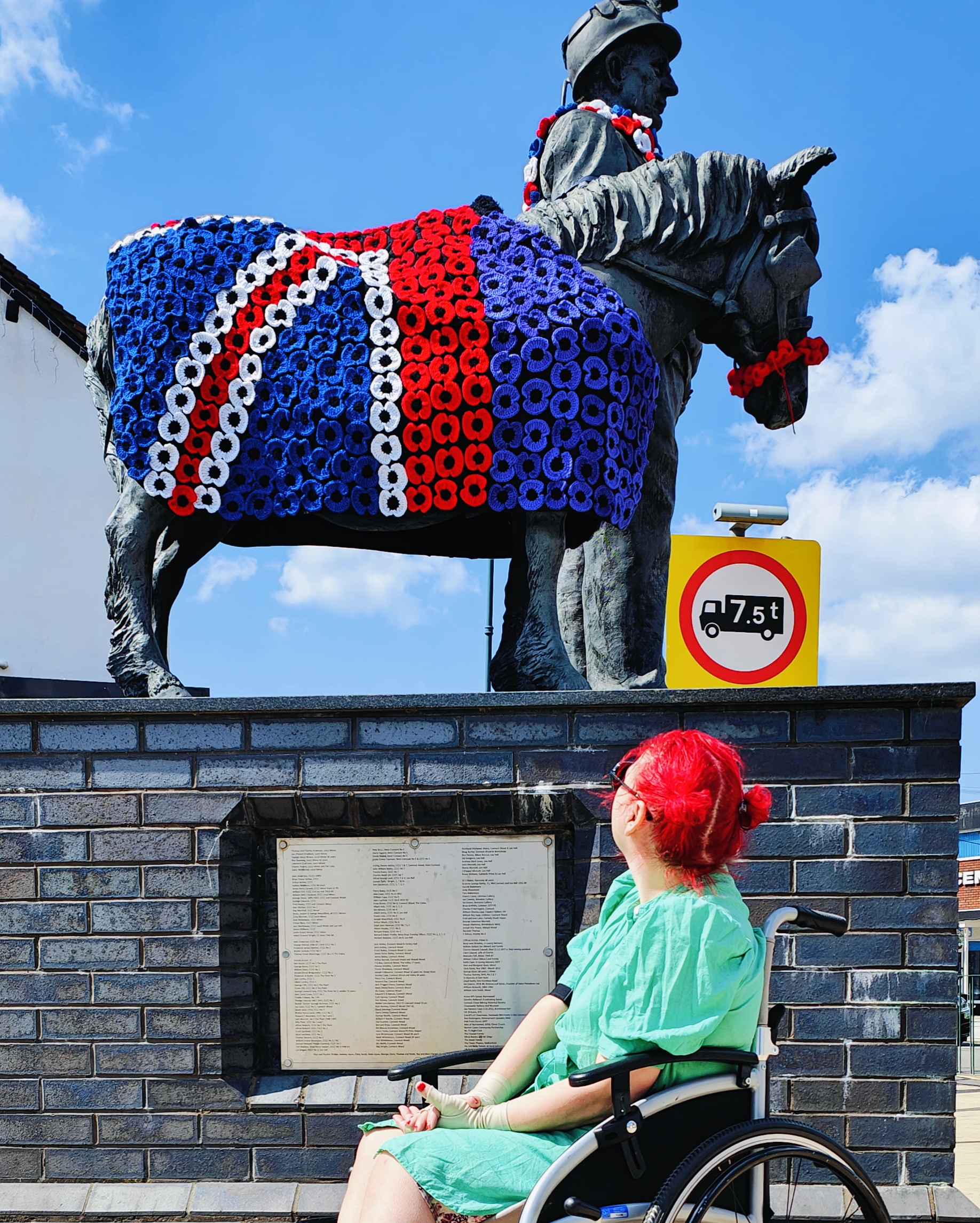 Me in my wheelchair wearing a bright mint green dress, with my red hair up in a bun, looking up at Scamp, the famous Burntwood Miner and Pit Pony statue. The statue is covered in crocheted poppies forming a Union Jack pattern, for the 80th anniversary of VE Day. I'm positioned by an information plaque on the brick wall base, with the statue towering above against a clear blue sky.