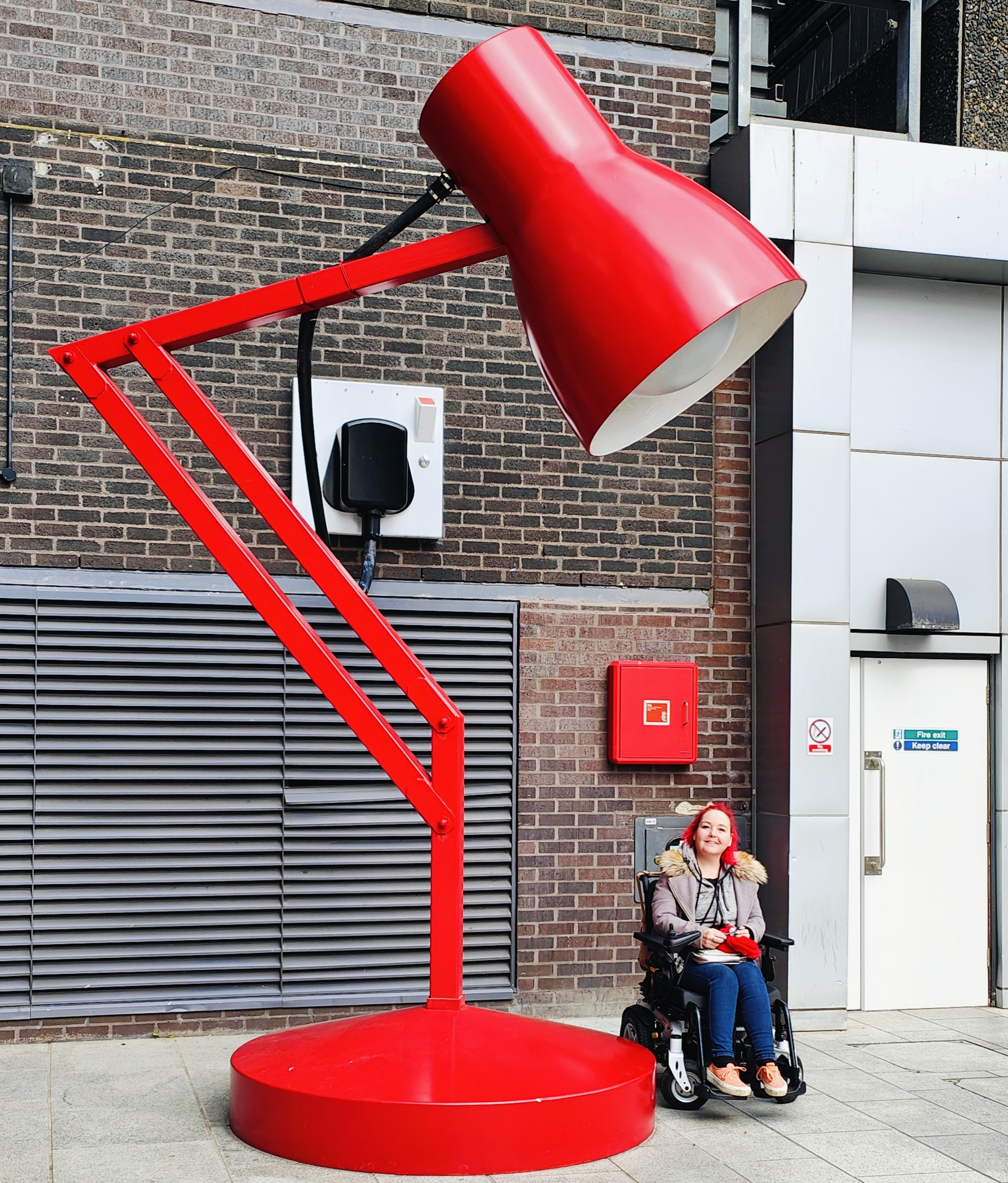 Me in my powered wheelchair next to a giant red Anglepoise-style lamp sculpture outside a modern building. I'm wearing a grey coat with fur-trimmed hood, jeans, and pink sneakers, with my signature bright red hair. I look tiny compared to the enormous lamp, which towers several metres high. The lamp and my hair are almost perfectly matched in their vibrant red colour against the brick and concrete background