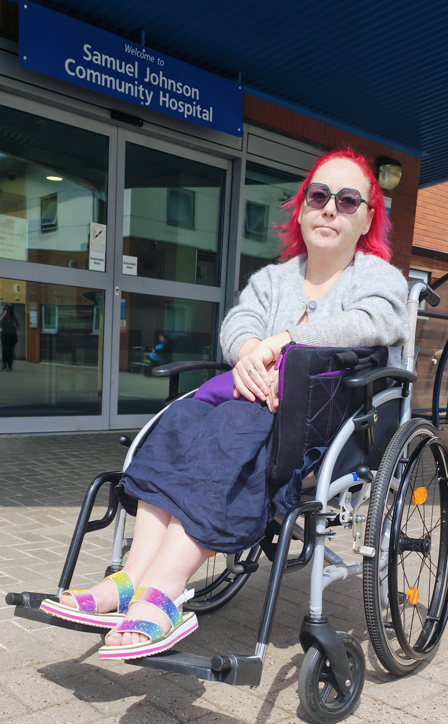 Me sitting in a manual wheelchair outside Samuel Johnson Community Hospital. I'm wearing a grey cardigan over a blue dress, with sparkly rainbow sandals adding a pop of colour. My bright red hair contrasts with the more subdued hospital entrance behind me. I'm looking directly at the camera with a slightly wry expression - the familiar 'hospital visit face' that balances between determination and resignation. The wheelchair is a standard manual model rather than my powered chair.