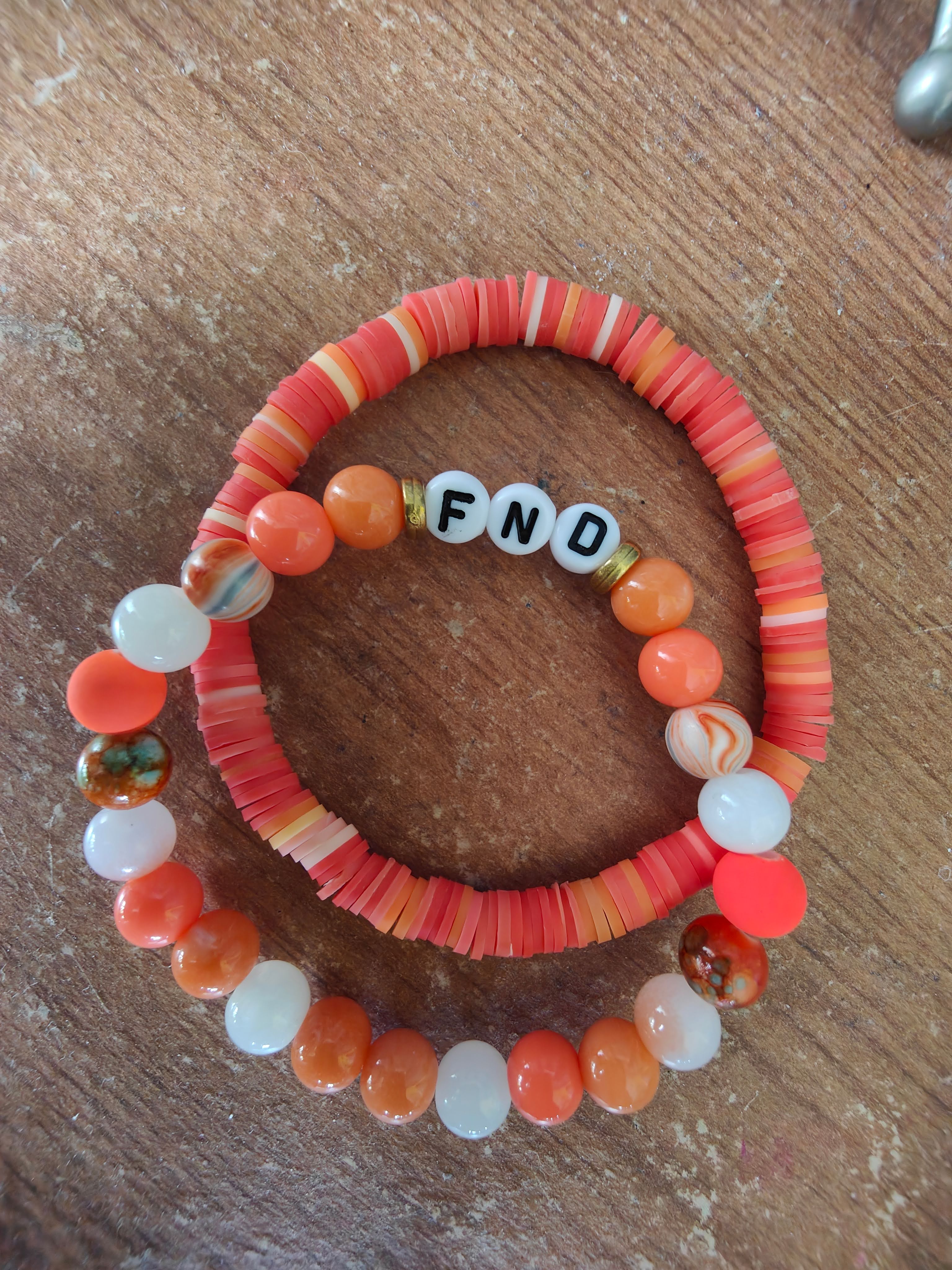 Two handmade bracelets arranged in a circle on a wooden surface. The bottom bracelet features alternating orange and white beads with a few marbled stones. The top bracelet has flat coral and peachy-orange disc beads with three white letter beads that spell 'FND' (Functional Neurological Disorder). Both bracelets share a similar warm colour palette of oranges, corals, and whites, creating a meaningful and stylish pair.
