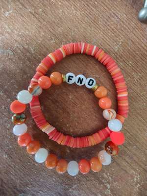 Two handmade bracelets arranged in a circle on a wooden surface. The bottom bracelet features alternating orange and white beads with a few marbled stones. The top bracelet has flat coral and peachy-orange disc beads with three white letter beads that spell 'FND' (Functional Neurological Disorder). Both bracelets share a similar warm colour palette of oranges, corals, and whites, creating a meaningful and stylish pair.