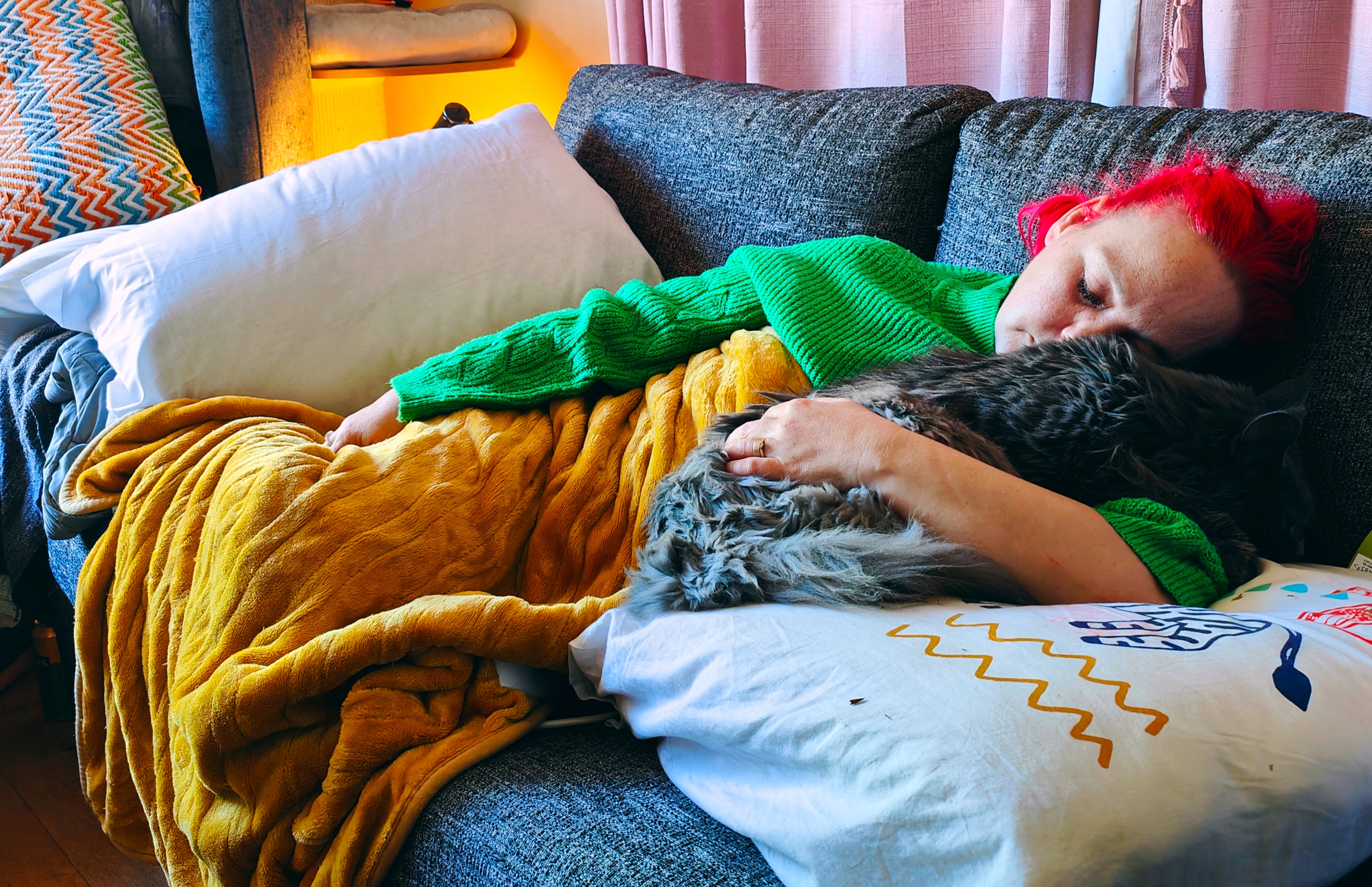 Me lying on the sofa with my bright red hair, wrapped in a cosy green jumper and mustard yellow blanket, cuddling with Mykavark, our fluffy Maine Coon cat. The sofa is covered with colourful cushions and throws in cheerful patterns. It's the perfect picture of a restful moment - the kind of therapeutic cat cuddle that makes everything feel a bit better.