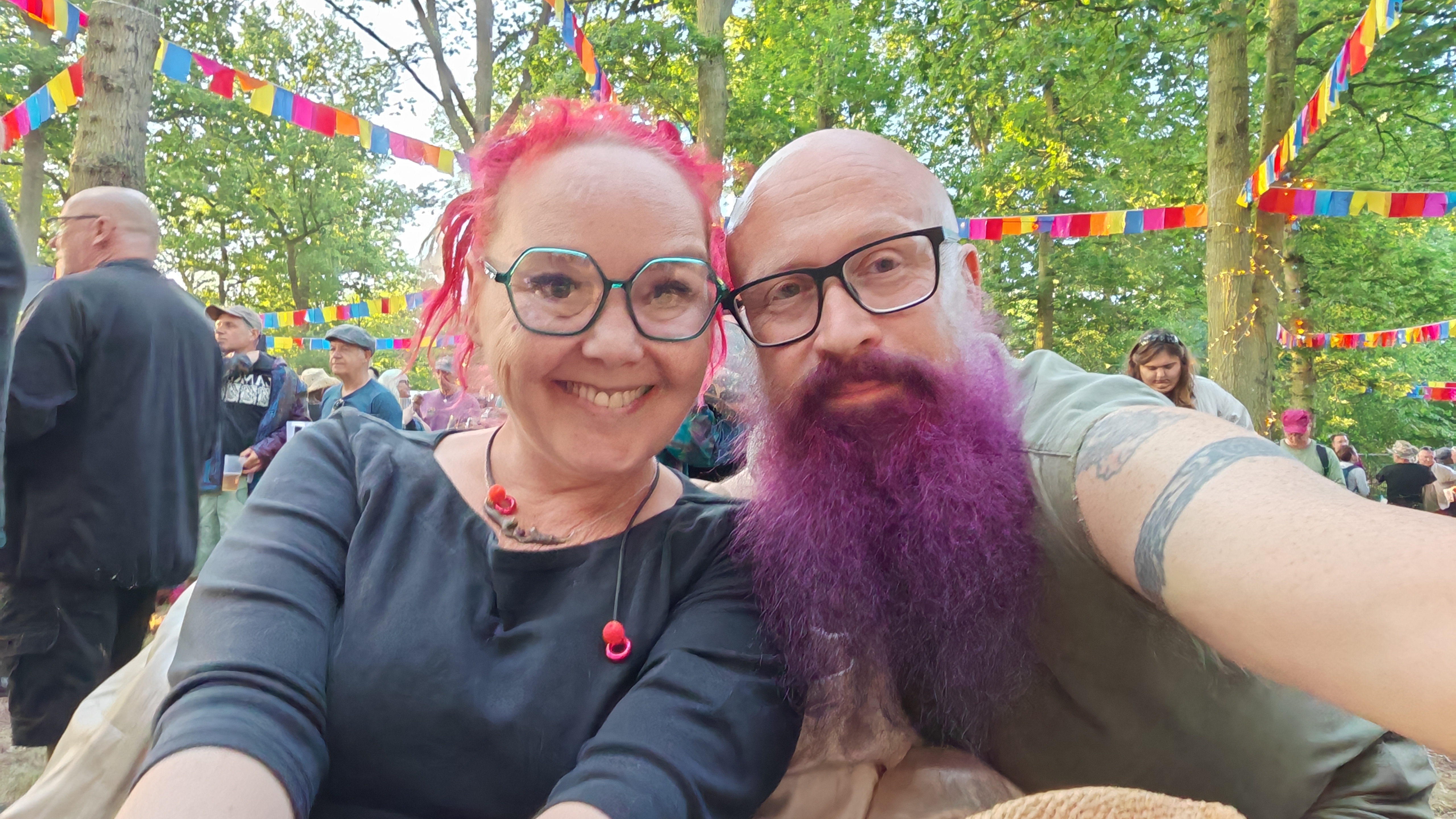 Sha and Paul at an outdoor festival, surrounded by colourful bunting. Sha has bright pink hair and is grinning, while Paul sports his magnificent purple beard. Both wearing glasses and looking far too pleased with themselves for two people who probably got lost trying to find the accessible toilets.