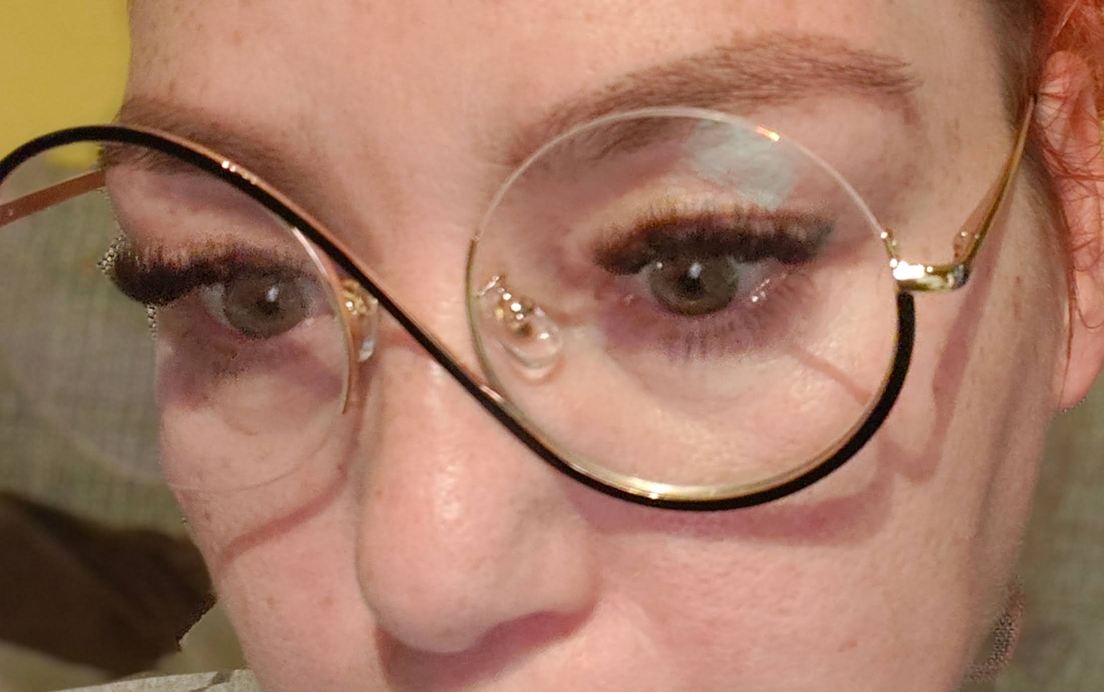 Close-up selfie of a woman with auburn hair wearing large round gold-rimmed glasses, showing off freshly done eyelash extensions and shaped eyebrows