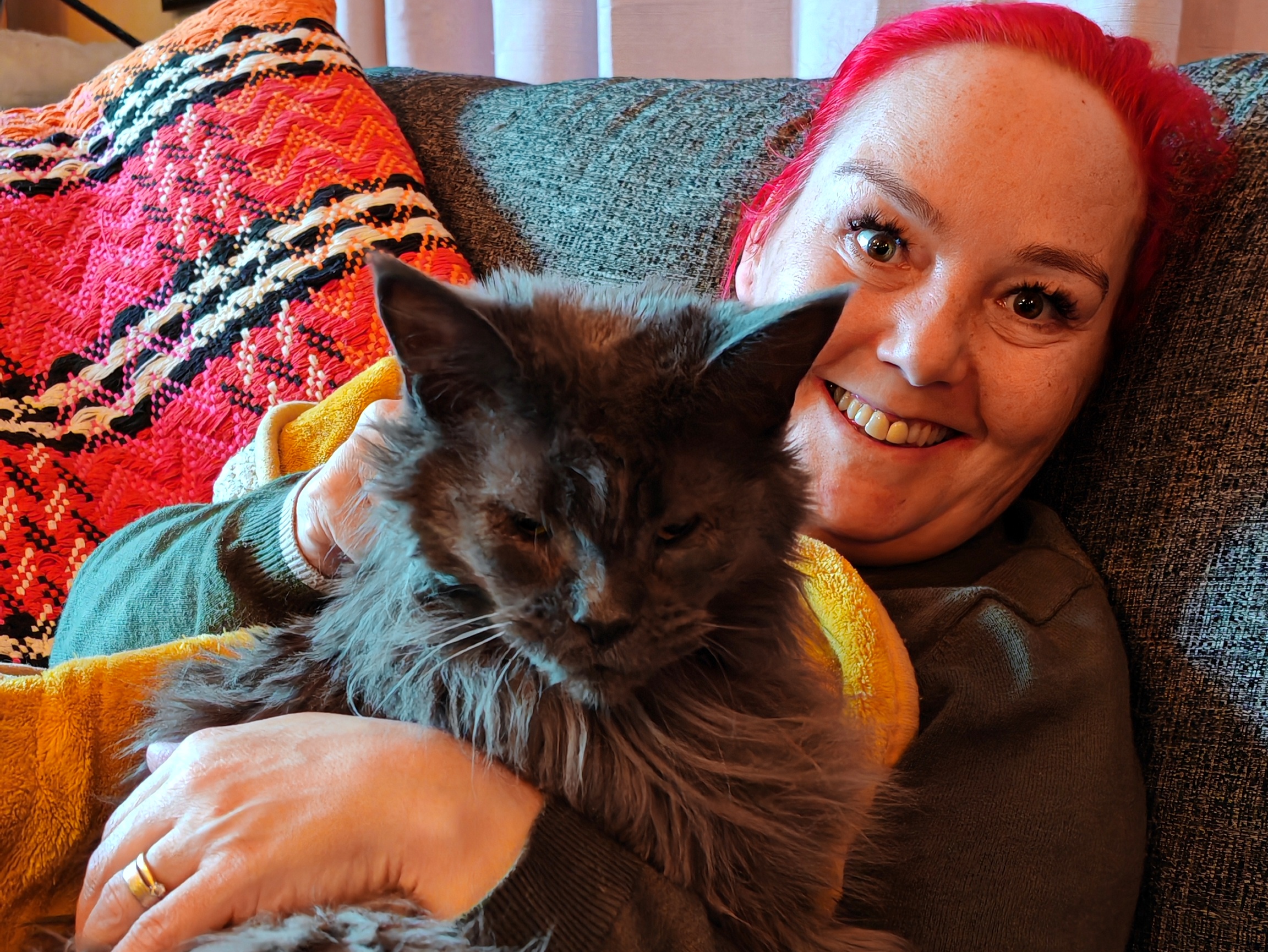 Me, a woman with bright red hair smiling warmly while cuddling Myka, a grey, fluffy, long-haired cat on a sofa with colourful patterned throws and blankets.