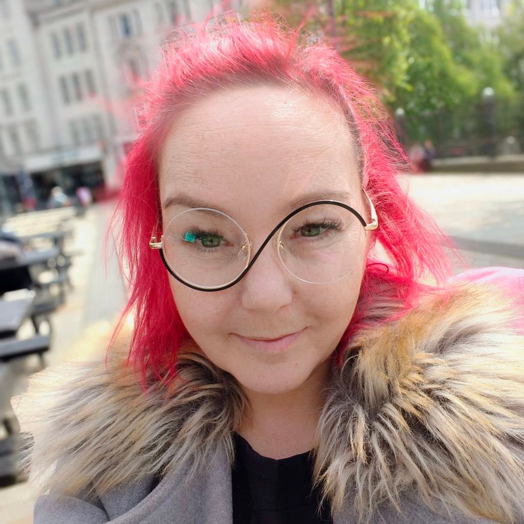 Close-up selfie of me with my bright pink hair (having a surprisingly good hair day!), round statement glasses, and a fabulous furry collar coat. I'm out and about in Lichfield town centre - you can see the blurred buildings behind me. Rocking my alternative look with a cheeky smile! My wheelchair isn't visible in this shot, but trust me, it's there and looking just as fabulous. Pink hair don't care - the wheels might slow me down, but the style never stops!