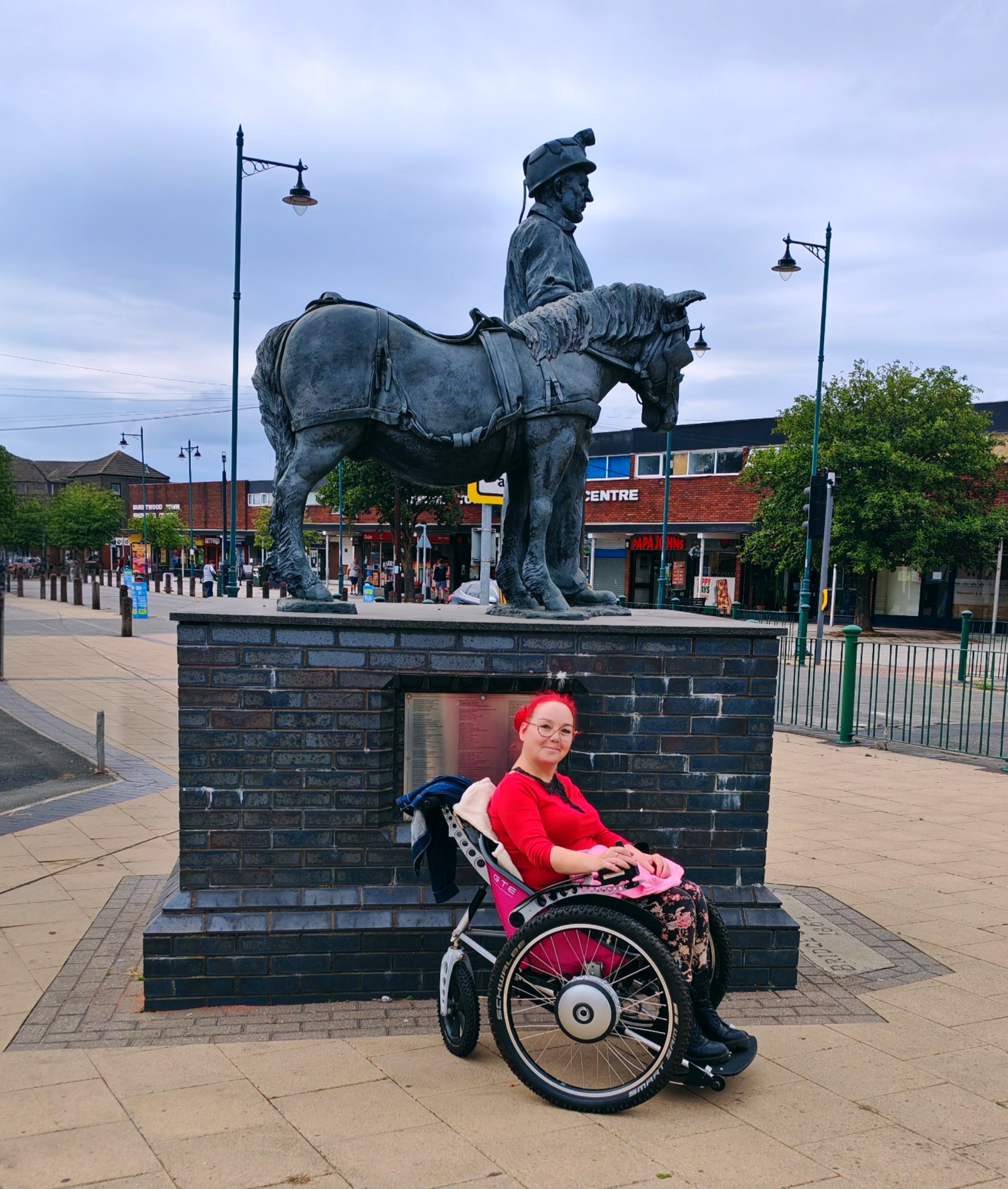 Sha, with bright red hair in a pink-seated Trekinetic GTE three-wheel wheelchair positioned in front of bronze miner and pit-pony statue in a town centre square, with shops and street lamps visible in background.