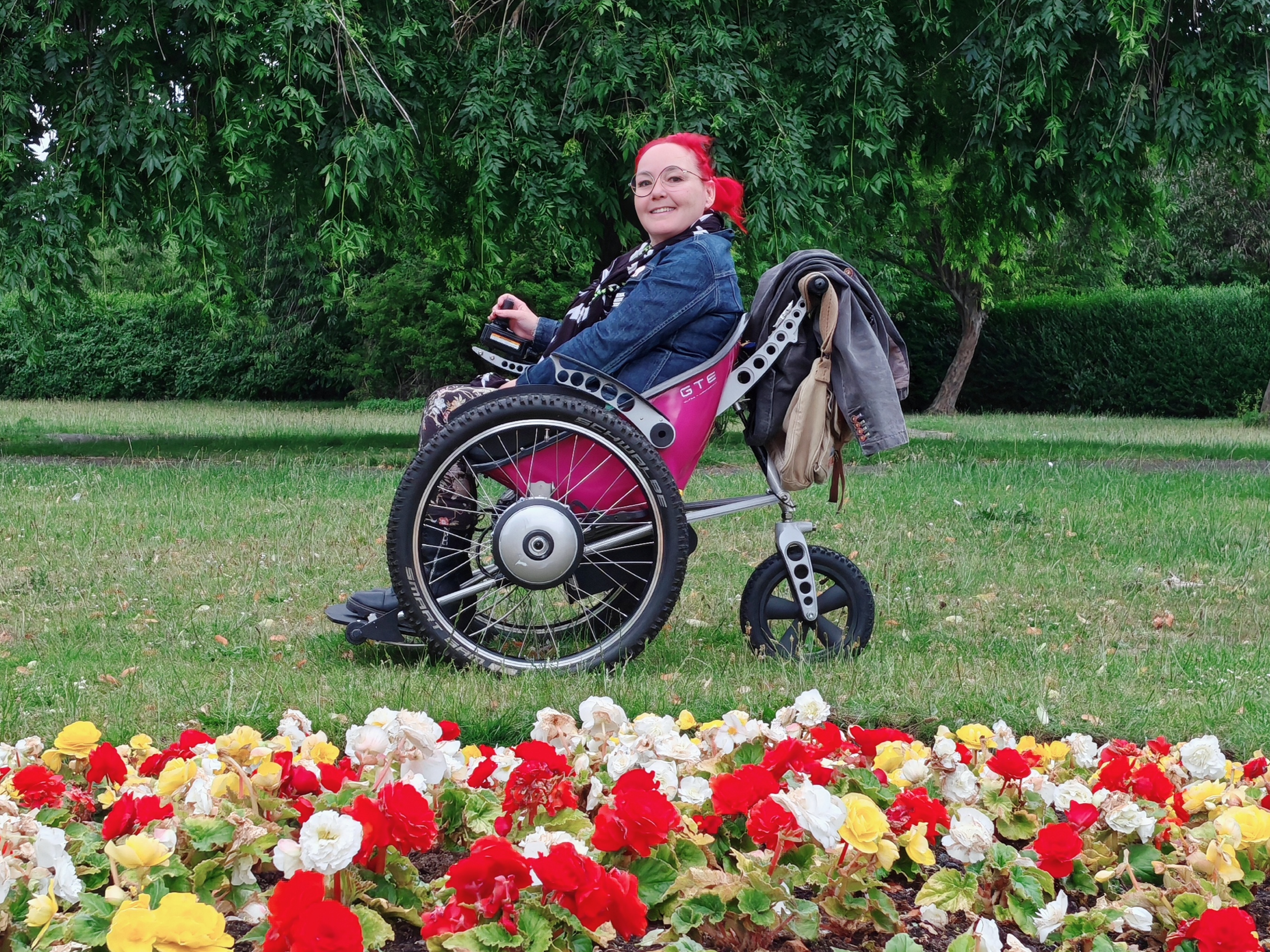 Sha with bright red hair in a pink-seated Trekinetic GTE wheelchair positioned on grass behind a colourful flower bed full of red, yellow and white begonias, with lush green trees in background.