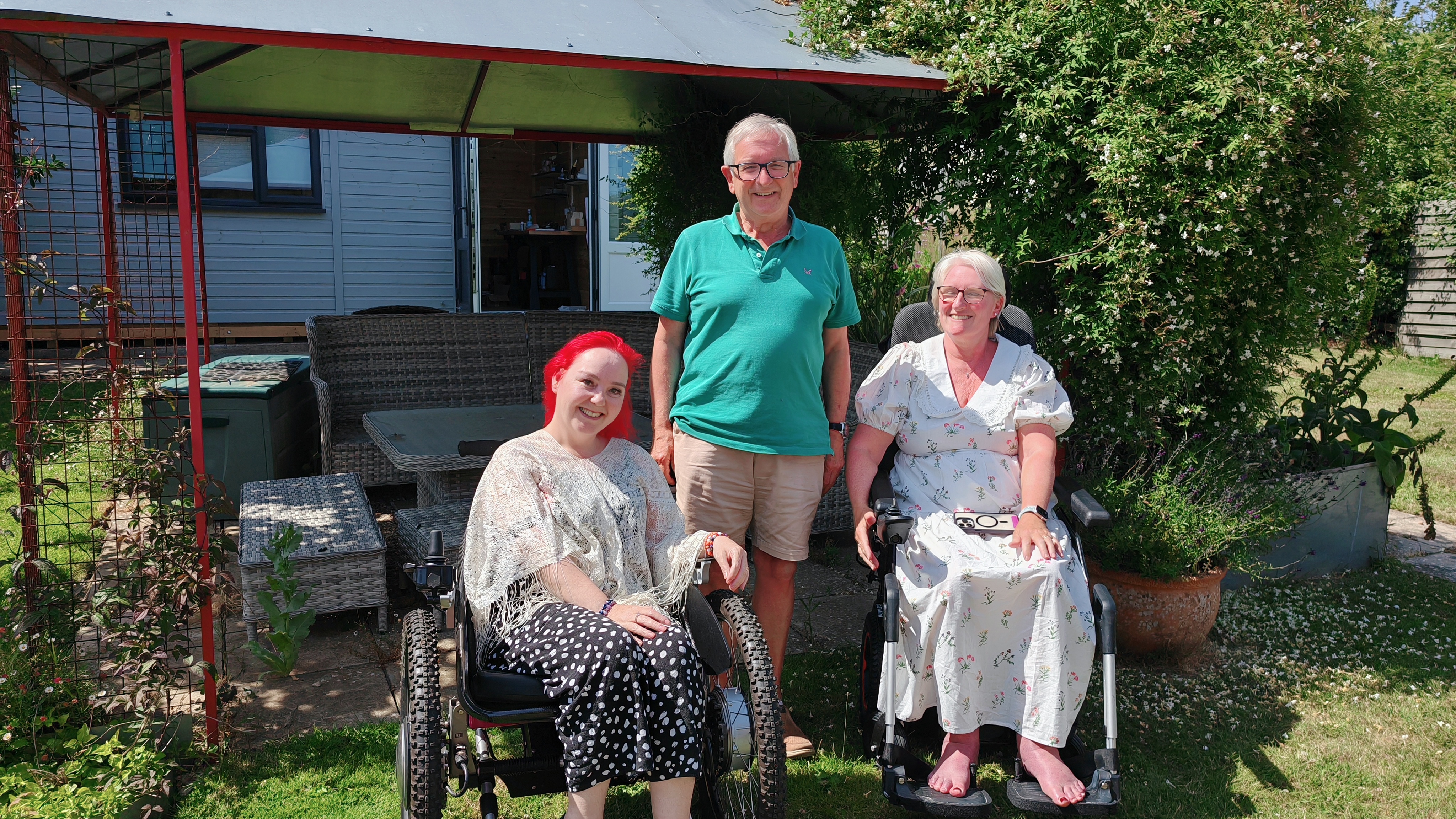 Three people enjoying a sunny garden moment - Sha with pink hair in the Trekinetic on the left, an older gentleman in a green polo standing in the centre, and another woman in wheelchair on the right, all posed happily in a garden with modern outdoor furniture.