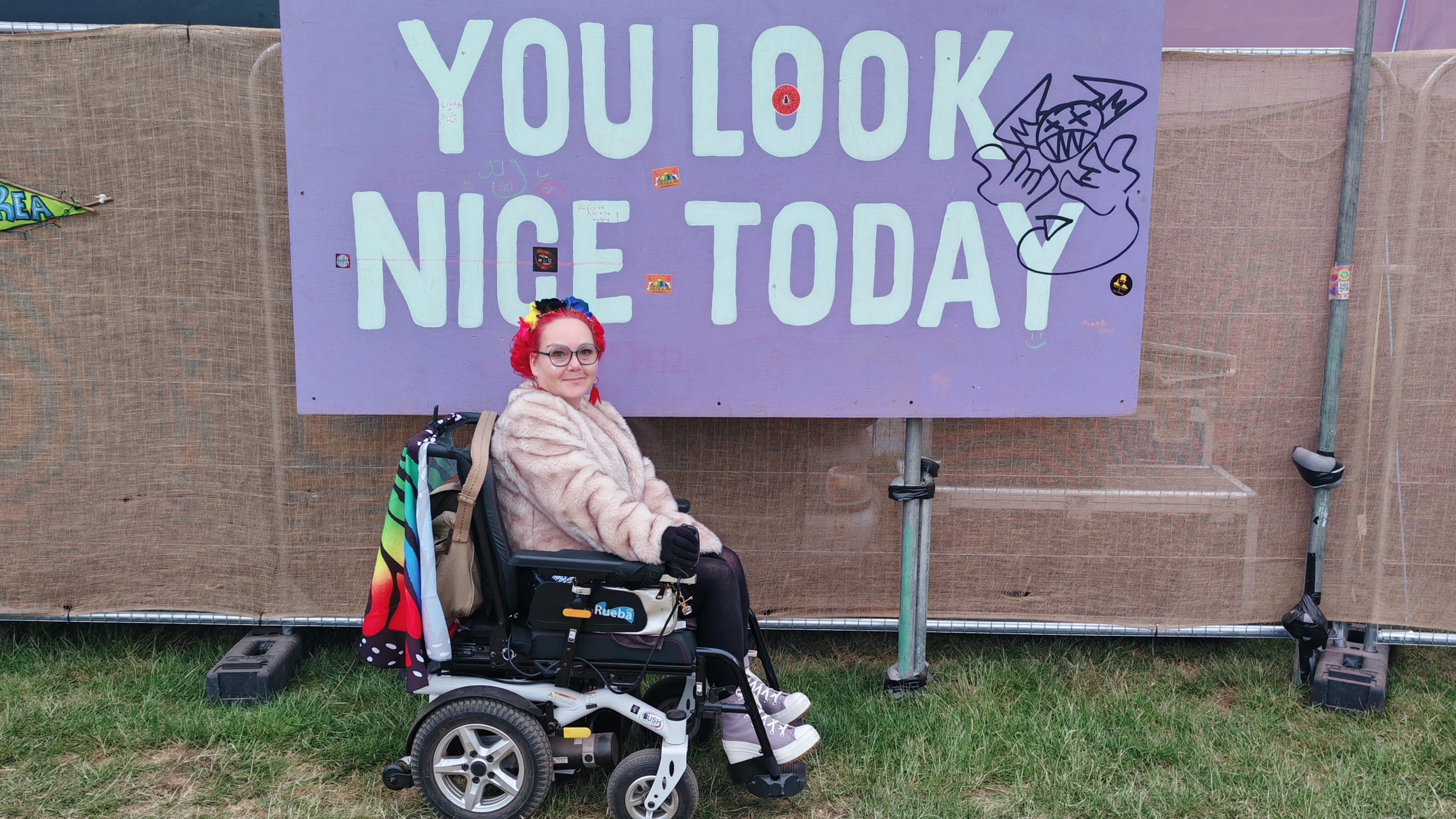 Woman with bright red hair and glasses sitting in a black and white power wheelchair, wearing cosy fluffy coat, positioned in front of large purple banner reading "YOU LOOK NICE TODAY" at outdoor festival with grass and temporary barriers visible. Perfect festival vibes! That banner is absolutely spot on - love how festivals just get the good energy flowing. The Rueba Rascal looks brilliant there too, proper festival-ready wheels! 💜