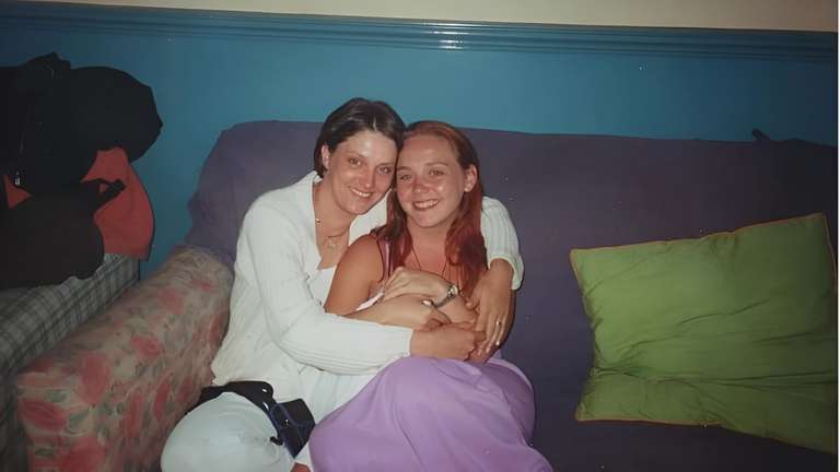 Two women sitting close together on a sofa, both smiling warmly at the camera. One woman wears a white top and the other wears a light-colored sleeveless top. The setting appears cozy and intimate, suggesting a close friendship moment.