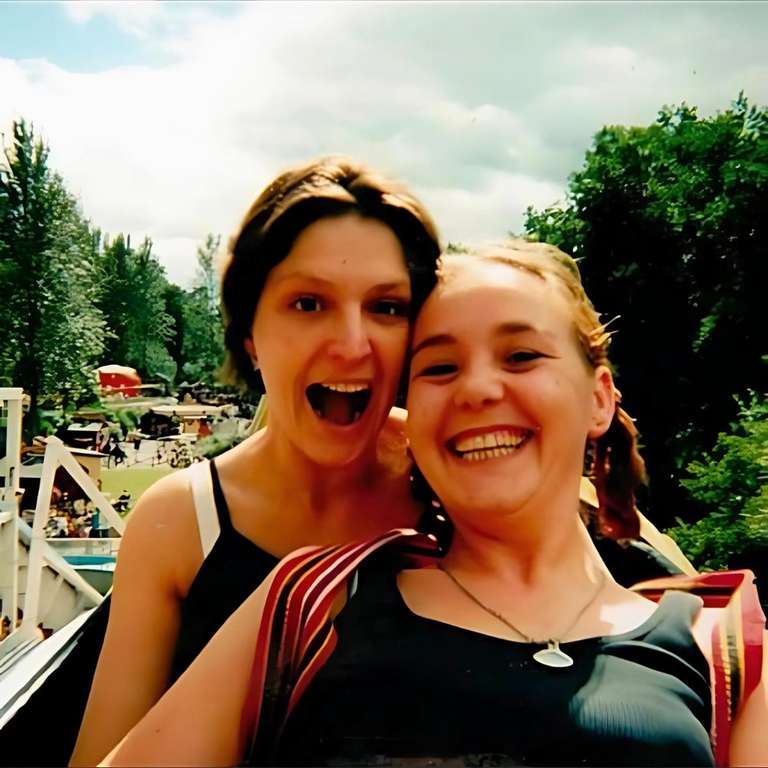 Two women on an amusement park ride, both laughing with pure joy. One has dark hair and wears a black tank top, the other has blonde hair. Trees and park structures are visible in the background, capturing a fun day out together.