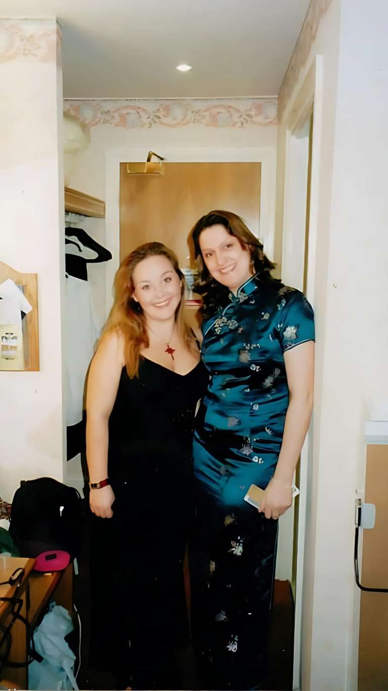 Two women standing together indoors, dressed up for what appears to be a special occasion. One wears a black dress with a red star necklace, the other wears a teal patterned Asian-style dress. Both are smiling and appear to be celebrating something together.