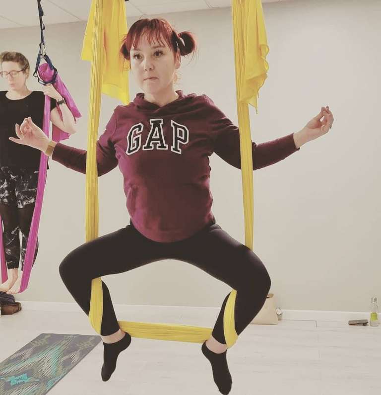 Woman with red hair in pigtails wearing a burgundy GAP hoodie and black leggings, suspended in bright yellow aerial yoga silks in a wide squat position with arms extended horizontally. She's in a bright white studio with other students visible in background, one using pink aerial silks. Woman has a focused, peaceful expression while demonstrating strength and balance in the aerial pose.