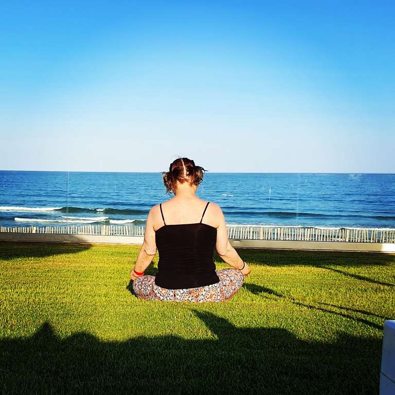 Woman with hair in a messy bun wearing a black strappy top and patterned leggings, sitting cross-legged in meditation pose on bright green manicured grass. Behind her is a stunning ocean view with deep blue water, white foam waves, and clear blue sky. A modern glass balustrade separates the lawn from the beach below. Golden afternoon sunlight creates dramatic shadows on the grass.