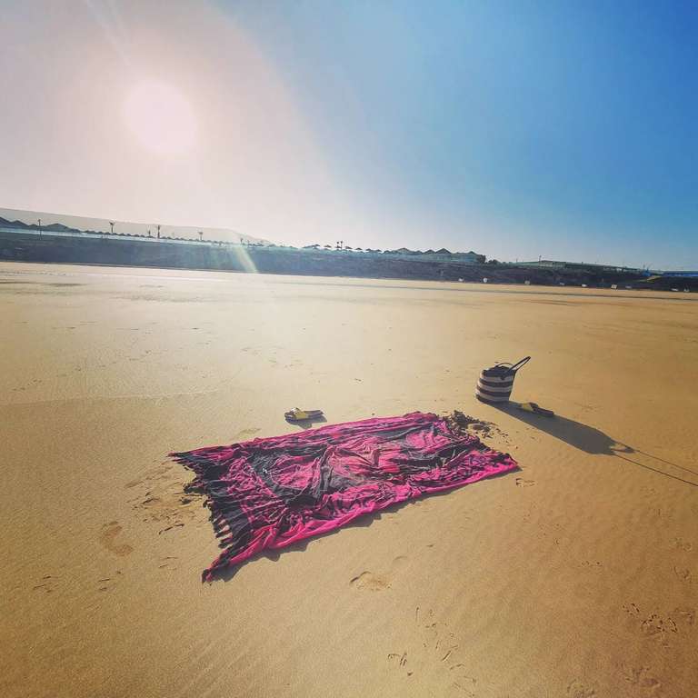 Empty beach scene with a vibrant pink and black patterned yoga mat spread on golden sand in foreground, alongside a striped beach bag. The expansive beach stretches into the distance under brilliant blue sky with wispy white clouds and bright sunburst. Rolling sand dunes and sparse coastal development visible on the horizon. The scene suggests a peaceful moment before or after yoga practice in this stunning Moroccan coastal setting.