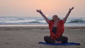 Woman in red patterned kimono-style cover-up over black top and leggings, sitting cross-legged on a blue yoga mat on sandy beach with both arms raised triumphantly toward a peach and pink sunset sky. The ocean waves are gently rolling behind her, and the wet sand reflects the warm sunset colours. Her face is tilted upward with a joyful expression of pure celebration and freedom.