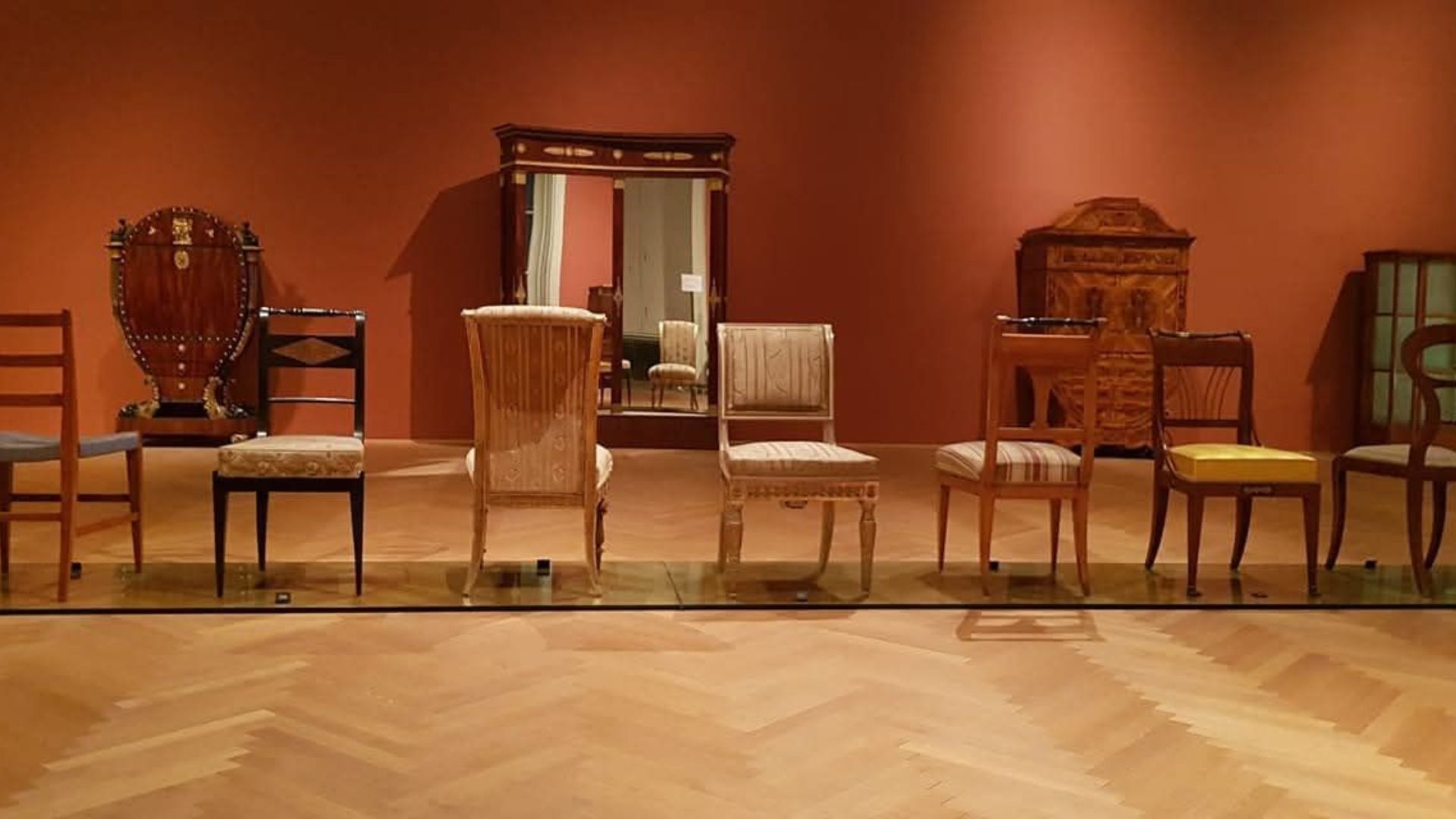 A museum display at Vienna's MAK (Museum for Applied Arts) showing a chronological arrangement of historical chairs against a warm terracotta-colored wall. The collection includes approximately eight chairs spanning different periods and styles, from simple wooden ladder-back chairs to more ornate upholstered pieces with decorative elements. Behind them stands a tall display cabinet with glass doors. The chairs are positioned on a polished parquet wood floor, and the museum lighting creates a warm, scholarly atmosphere typical of design history exhibitions.