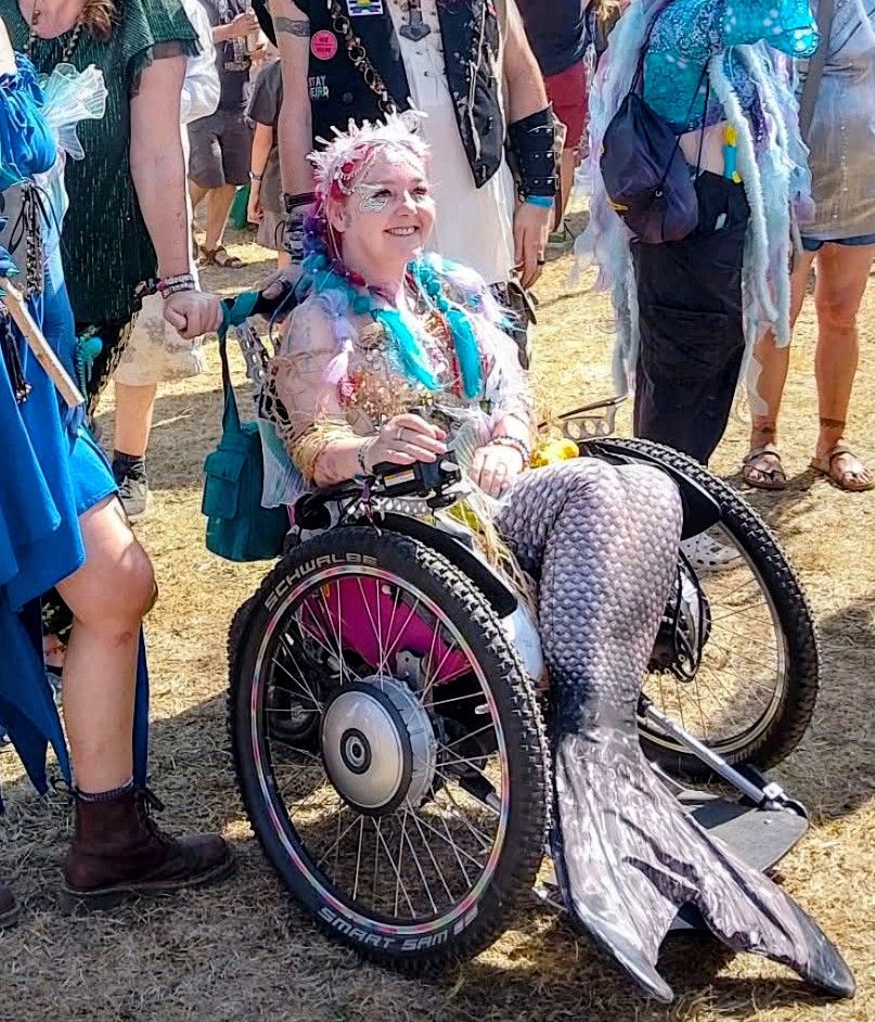 A woman in a wheelchair at Beautiful Days festival wearing an elaborate mermaid costume with iridescent silver and blue scales, flowing fabric tail, and face paint with decorative elements in her hair. She's sitting in a three-wheeled Trekinetic powered wheelchair with distinctive large wheels, surrounded by other festival-goers also in colorful costumes. The scene is on dusty festival ground, capturing a joyful moment of festival accessibility and inclusion, showing that wheelchair users can fully participate in the festival experience and creative costume culture.