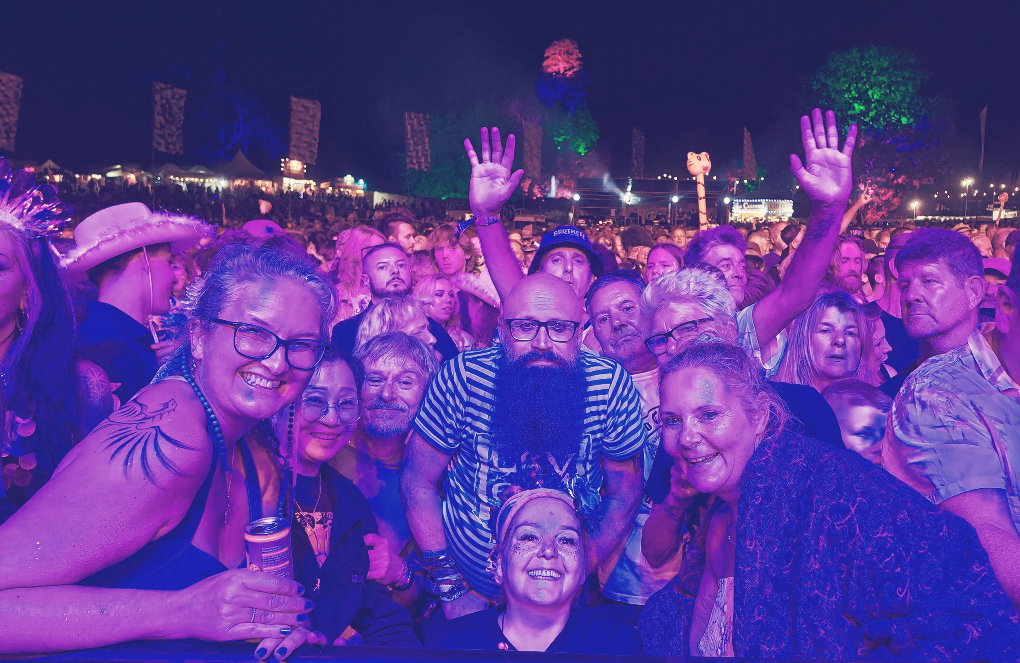 A crowded festival scene at Beautiful Days with festival-goers packed together at the barrier in front of the main stage under vibrant purple and pink stage lighting. The crowd includes people of various ages celebrating with hands raised in the air, dancing and singing along. In the foreground, a group including a man with a distinctive purple beard and glasses can be seen among the enthusiastic crowd. The atmosphere captures the energy and joy of a live music performance with decorative banners and festival infrastructure visible in the background.