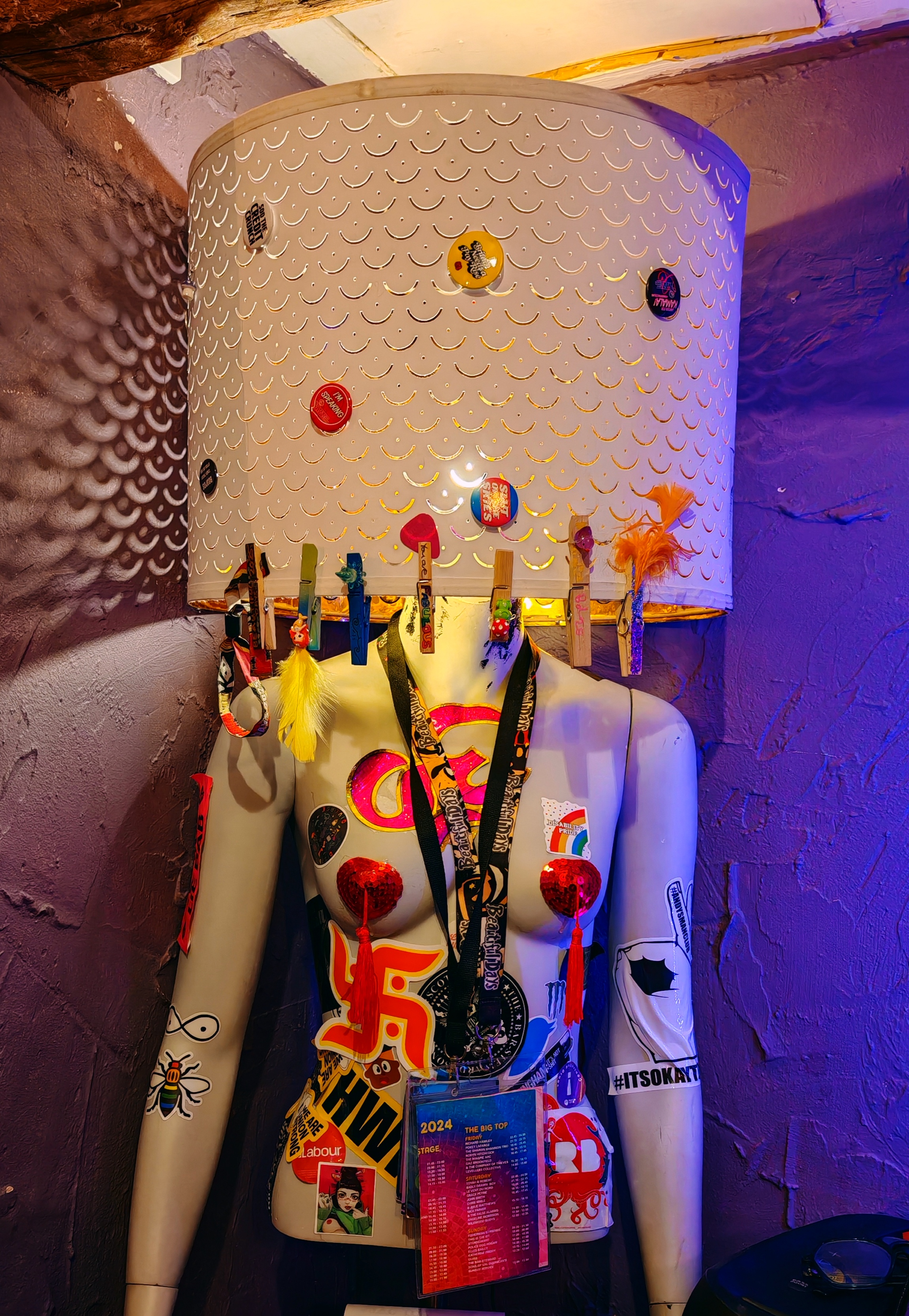 A half-mannequin torso with a white lampshade featuring a fish scale pattern in place of a head, displayed against purple-lit stone walls. The mannequin is covered with numerous colorful festival badges, pins, lanyards, and commemorative items collected from various music festivals and events. The items include wristbands, small decorative pins, festival passes, and other memorabilia attached to the mannequin's arms and torso, creating a vibrant display of festival culture and collecting traditions.