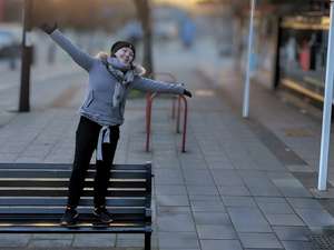 Sha, a woman in her early 50s, standing confidently on a bench at Sankey's Corner in Burntwood, arms spread wide in a joyful pose, wearing a grey jacket, scarf and beanie, with a big smile on her face - capturing a moment of freedom and happiness before FND and CFS changed her mobility.