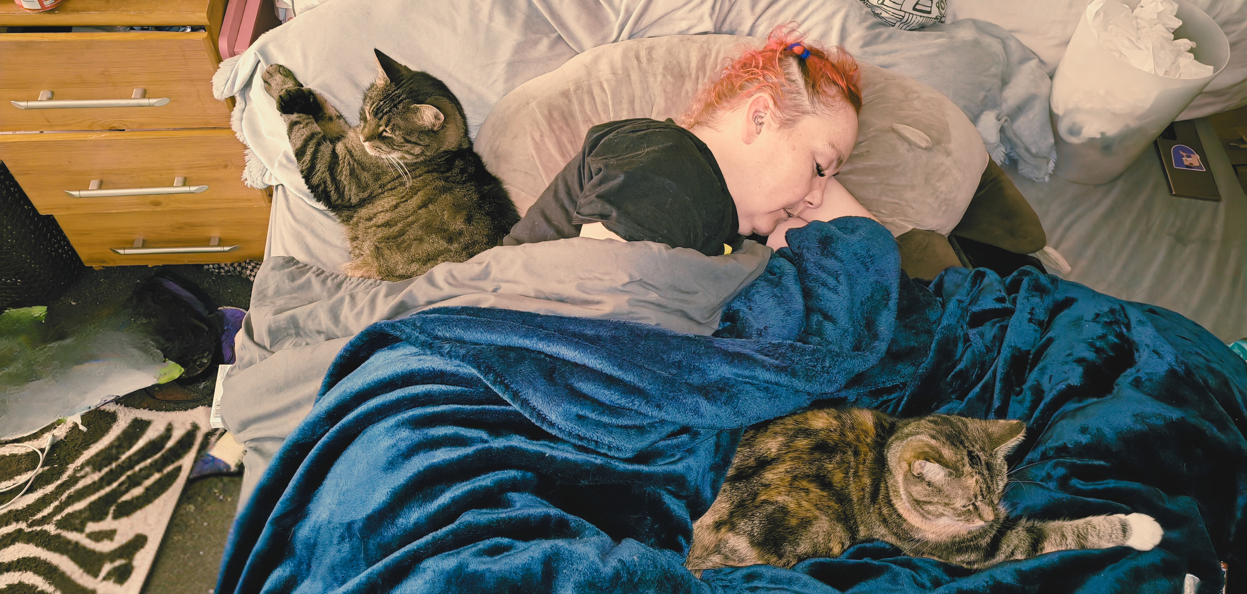 A woman with vibrant orange-red hair rests in bed under a teal blanket, flanked by two tabby cats who've appointed themselves as furry nurse companions during her bout with COVID - one perched on the pillow above, the other stretched out contentedly on the blanket beside her.