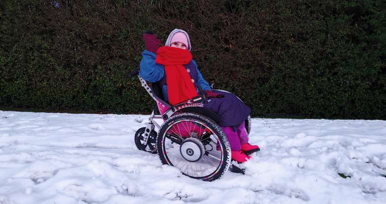 Sha sits in her pink and black Trekinetic wheelchair on a snowy grass verge, wearing a pink knitted hat, bright red scarf wrapped around her face, blue jacket, purple trousers, and pink boots. She's giving a wave with one hand while bundled against the cold. The wheelchair's large wheels have snow on them, and there's a hedge in the background.