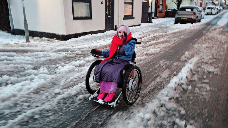 Motion-blurred photo of Sha rolling down a snowy street in her Trekinetic wheelchair, pink hat and red scarf visible as she moves. Snow is piled high on the pavement to her left, with houses and parked cars visible in the winter scene. The motion blur captures the speed and momentum of her movement through the snowy conditions.