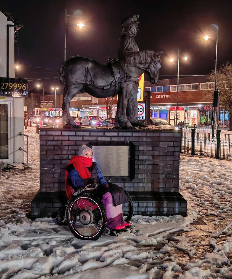 Night-time photo of Sha in her Trekinetic wheelchair positioned in front of the Burntwood Miner statue - a large bronze sculpture of a miner with a pit pony mounted on a brick plinth. Sha is bundled in her winter layers with pink hat and red scarf, her wheelchair visible against the snowy ground. Street lights illuminate the scene with a warm glow, and shops can be seen in the background.