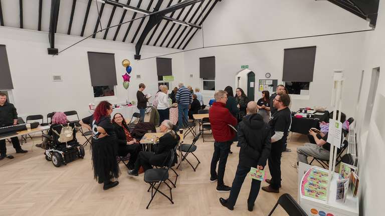 nside Chase Terrace Community Centre during the Creative Burntwood launch - a proper busy community hall with gorgeous high ceilings and exposed beams. People are milling about, chatting, and doing that lovely 'peopling' thing. You can spot yours truly in my wheelchair, pink hair and all, sitting at a table. There are balloons, folding chairs, and that brilliant buzz of community happening. This is what I overdid it for, and honestly? Worth every spoon I spent (even if the Ellyllons are making me pay for it today).