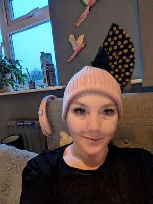 Sha smiling at the camera wearing a cosy peach-coloured beanie hat, seated on her sofa at home with decorative birds on the grey wall behind her and a "Severe delays" sign visible on the radiator behind her