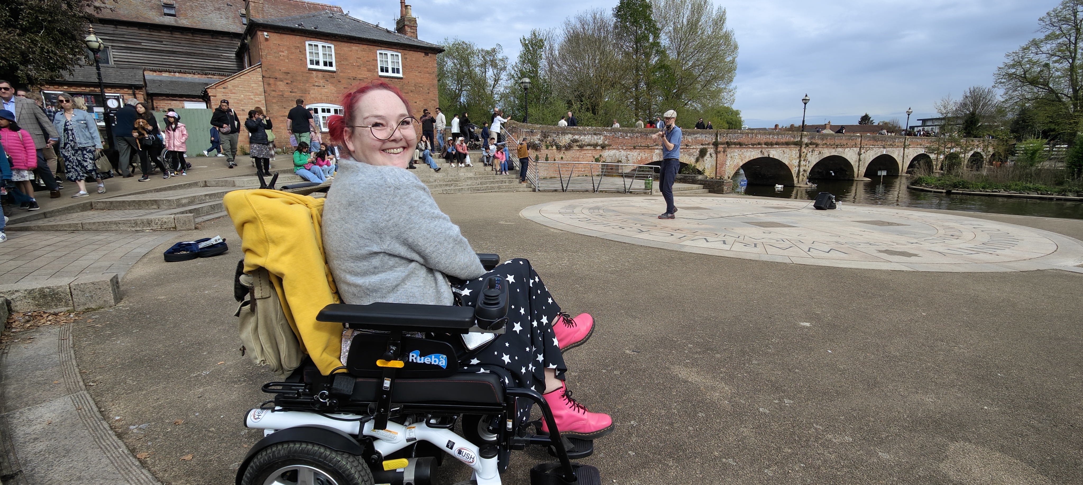My wife beaming with pink hair, round glasses and a cheeky grin as she sits in her wheelchair at Stratford-upon-Avon riverside. Behind her, a violinist busker adds a bit of culture to the scene while looking like he's attempting to serenade the swans under the iconic red brick bridge. Her star-patterned trousers and bright pink Doc Martens prove that disability doesn't mean sacrificing style. Just another day in Shakespeare country after a proper gig.