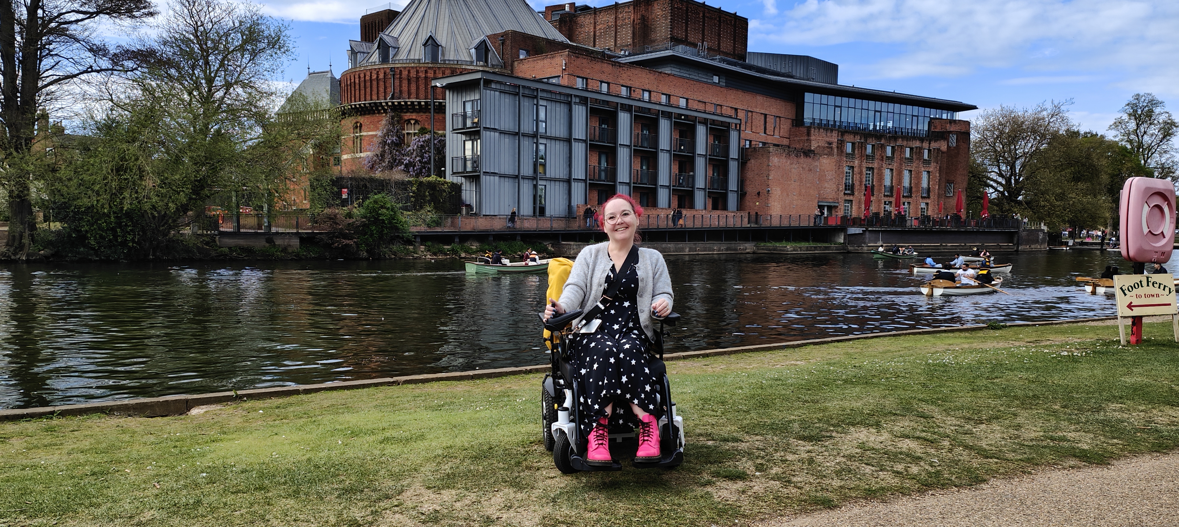 My wife showing off her pink hair and stylish star-patterned dress by the River Avon, with the magnificent brick and glass faΓ§ade of The Swan Theatre behind her. Several boats drift by on the water as tourists row past the theatre in what looks like a daft attempt to get cultured by osmosis. There's a sign for a 'Foot Ferry to town' nearby - because nothing says 'Shakespeare's England' like avoiding a five-minute walk by paying for a boat ride. A rare bit of spring sunshine has graced Stratford for our post-gig wander.