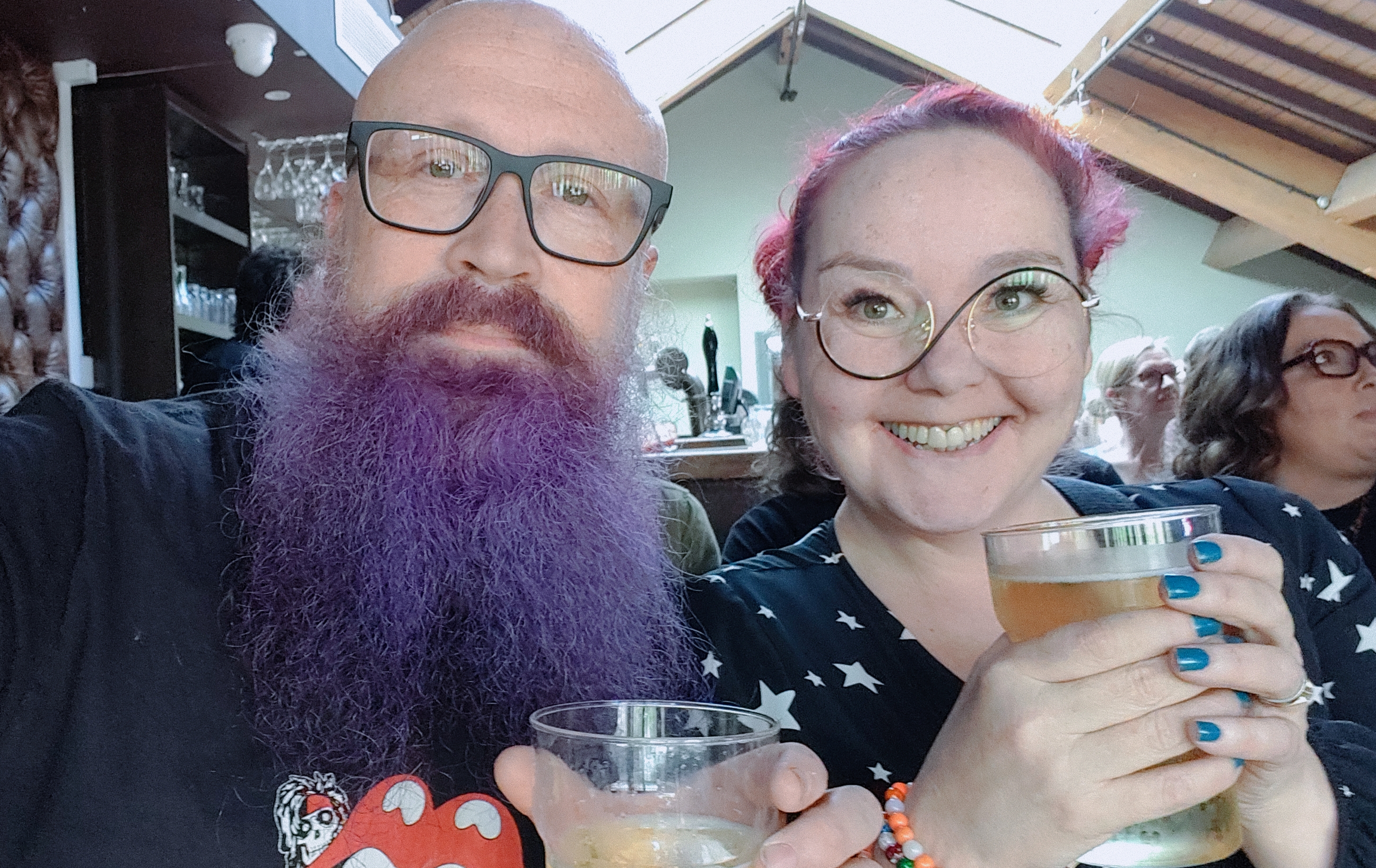 Selfie at a pub with my magnificent purple beard stealing the show and my wife sporting vibrant pink hair and round glasses, both grinning like we've just heard the world's most British joke. We're holding drinks at Cox's Yard in Stratford-upon-Avon before a Miles Hunt gig. Because nothing says 'responsible middle-aged couple' quite like colourful hair and beer on a weekend afternoon..