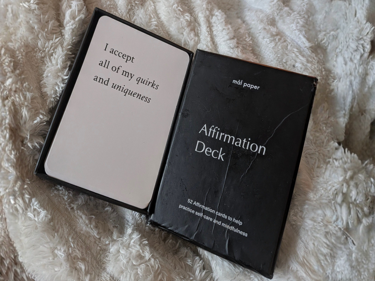 Here's the alt text:  **Alt text:** *A Mål Paper Affirmation Deck box sits open on a soft cream fluffy blanket. The card on top reads "I accept all of my quirks and uniqueness" in elegant italic script. The black box lid shows the deck title and the words "52 affirmation cards to help practice self-care and mindfulness."*
