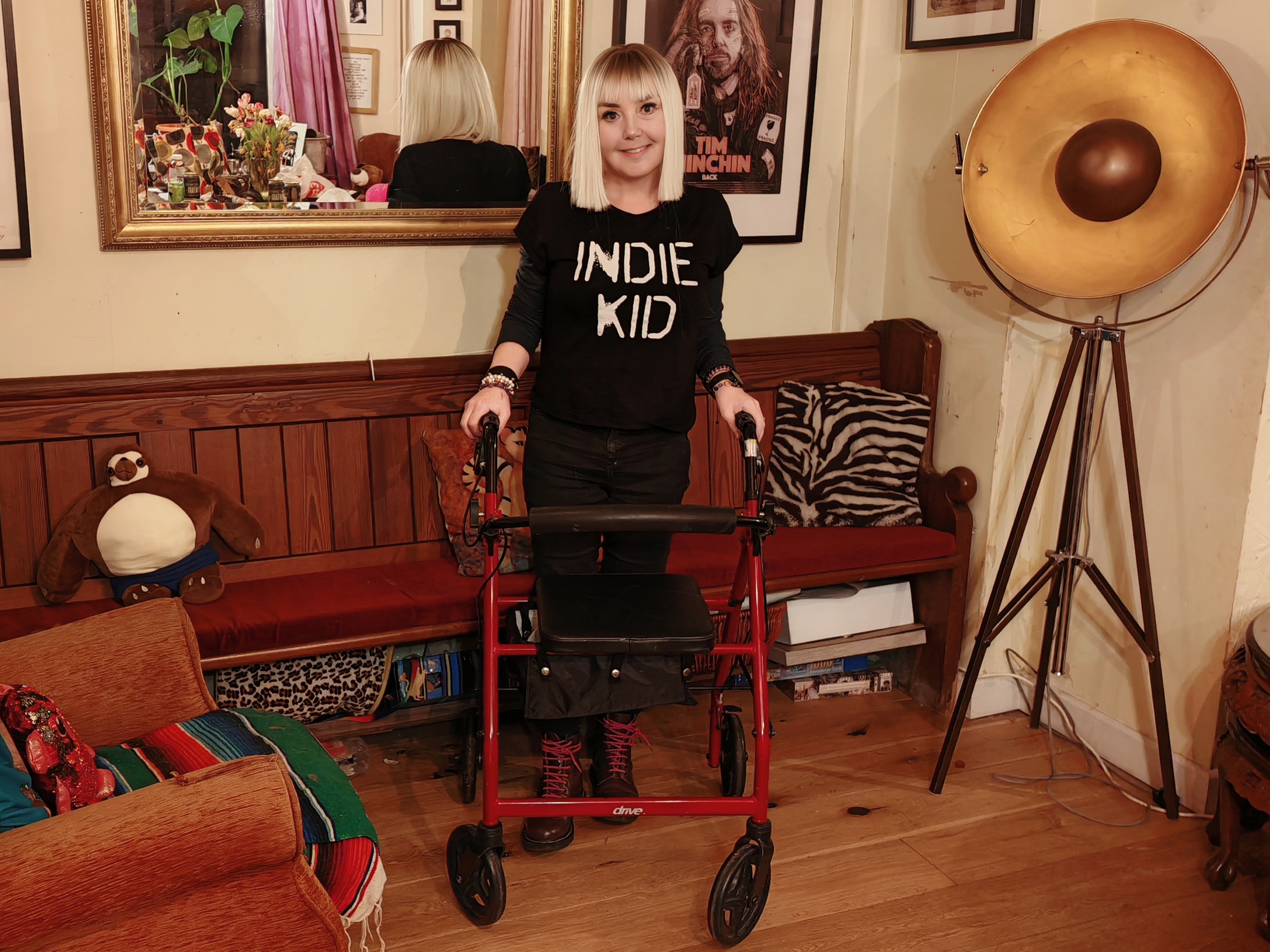 Sha, a woman in her early 50s with a platinum blonde bob, stands smiling in her living room holding a red rollator walker. She's wearing a black "Indie Kid" sweatshirt, black jeans, and pink-laced boots. The room behind her is eclectic and characterful — a church pew, a giant studio lamp, zebra print cushions and a Tim Minchin poster on the wall. Exactly the home of someone who has made a conscious decision to grow old disgracefully.