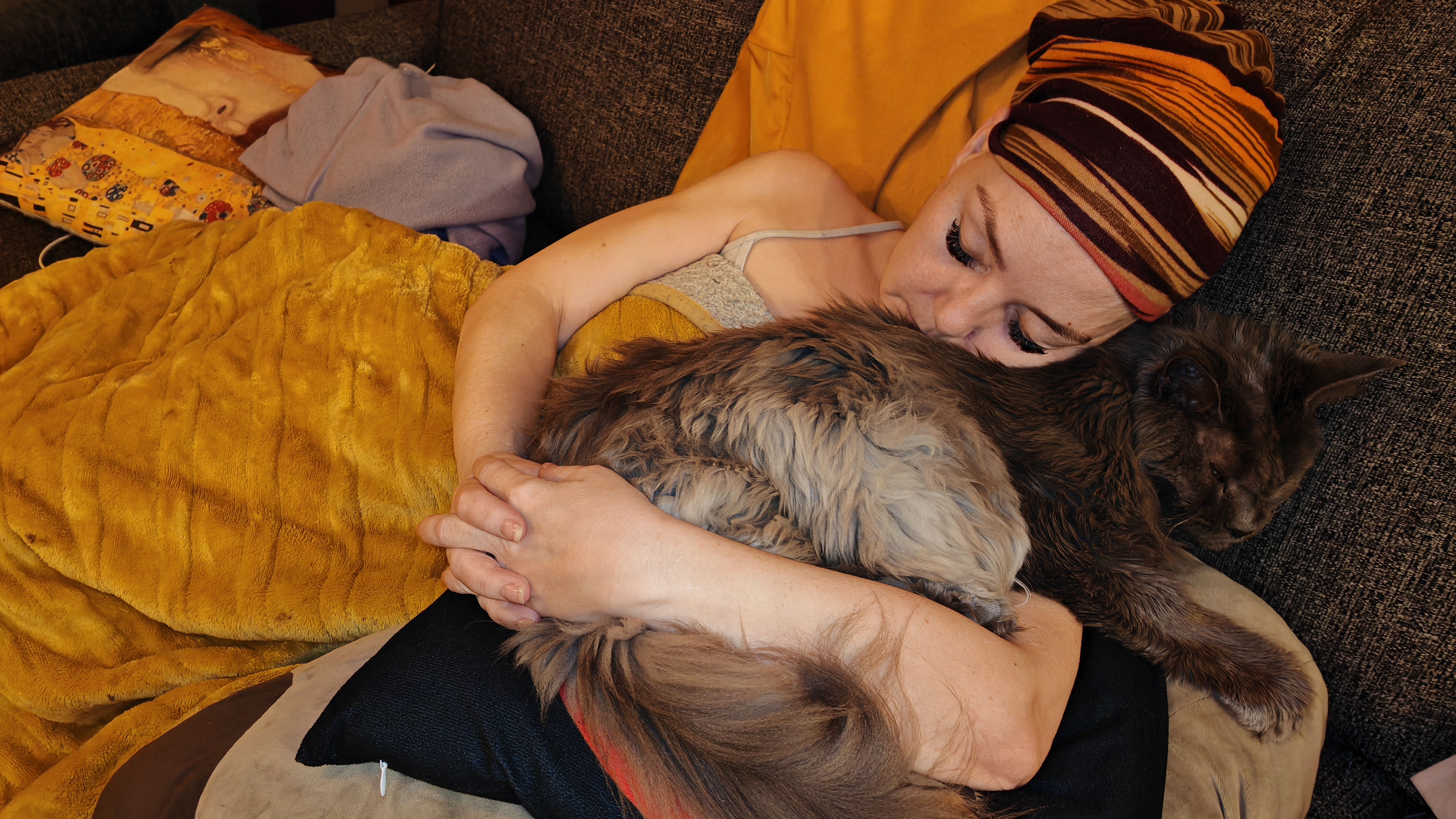 Photograph taken from above of MrsVark lying on a grey sofa, wrapped in a mustard yellow fleece blanket, cuddling MykaVark, a grey Maine Coon cat. She's wearing a striped headwrap in brown, orange, and cream tones, and is gazing down at Jasper with a soft, affectionate expression. Myka looks entirely unbothered by the whole arrangement, as is traditional.