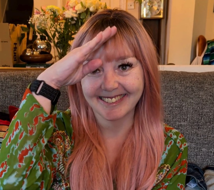 Sha, a woman with long pink hair and a big grin, giving a slightly chaotic salute to the camera. She's wearing a gorgeous green and orange patterned top and sitting on the sofa like the absolute trooper she is. Yes, she did manage to get dressed today. Yes, she is very proud of herself.