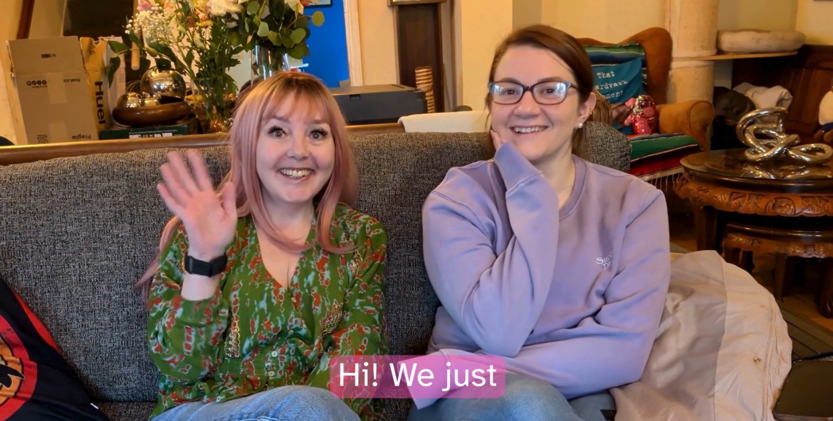 Two women sitting together on a grey sofa, mid-conversation and grinning. On the left, a woman with pink hair in a green floral blouse is mid-wave at the camera — that's me, your Mrsvark, having a reasonably good spoon day by the looks of things. On the right, a woman with dark hair pulled back, glasses, and a lilac sweatshirt, smiling in that way that suggests she's about thirty seconds from saying something dry — that's Aimee, the Aimeevark herself. Behind us: flowers, general organised chaos, and if you look carefully, a cushion that says "That Aardvark" something. Because of course it does. 🐾💜