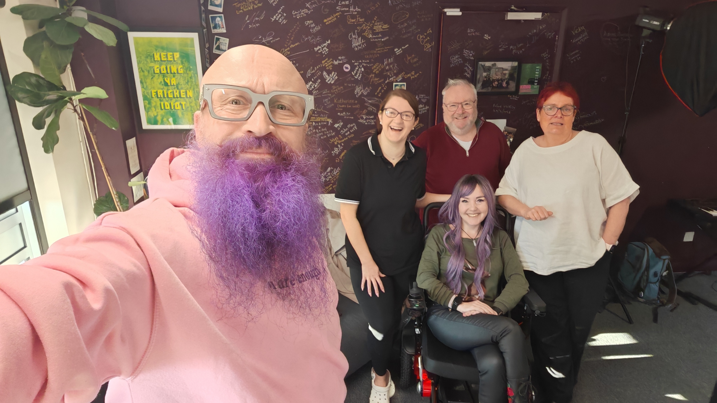 A selfie taken at Lichfield Radio studio. In the foreground, a bald man with a magnificent purple beard and pink hoodie grins at the camera — that's our #TechGuyPaul. Behind him, five people smile and laugh in front of a dark wall covered in signatures. Centre front is a young woman with gorgeous purple hair seated in a power wheelchair — Aimeevark in her natural habitat. Behind her, Ruth Redgate beaming on the left, Paul's co-host in the middle, and Sha on the right looking like she hasn't been up since 6am (she has). The poster on the wall behind them reads "KEEP GOING YA FRIGGEN IDIOT" — honestly the most appropriate studio decor imaginable. 💜🎙️