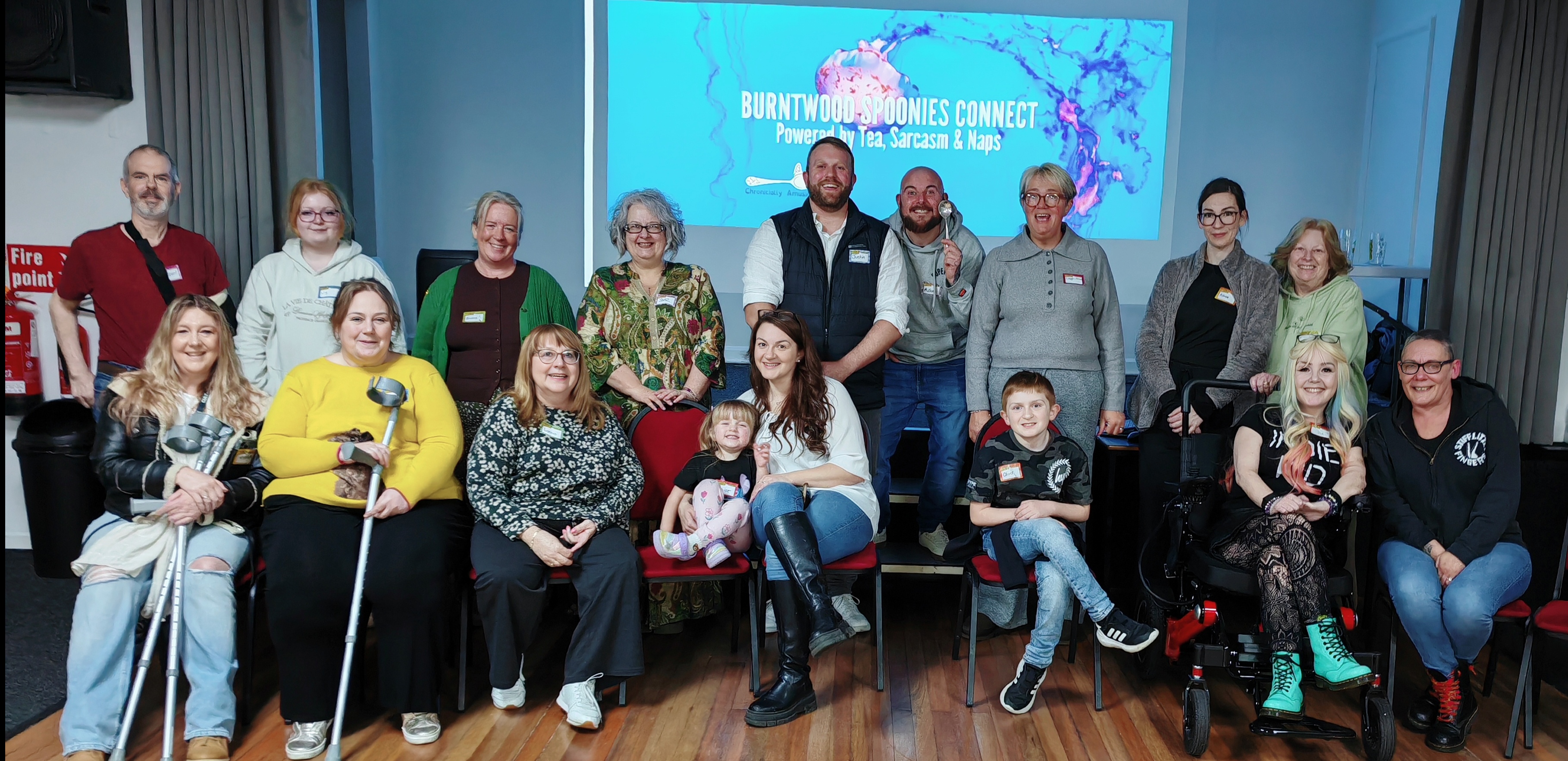 A group of around 18 people — spoonies, carers, and a couple of very welcome small humans — gathered together and smiling for a group photo at the first Burntwood Spoonies Gathering at The Grangemoor, 24th March 2026. The room is warm and relaxed; people are seated and standing in two rows, name badges on, genuinely happy faces all round. Mobility aids are visible and completely unremarkable — crutches in the foreground, a powerchair on the right. Behind the group, a large projected screen displays the Burntwood Spoonies Connect branding: "Powered by Tea, Sarcasm and Naps." One attendee is holding a giant novelty spoon aloft, because of course he is. This is what community looks like. 💜